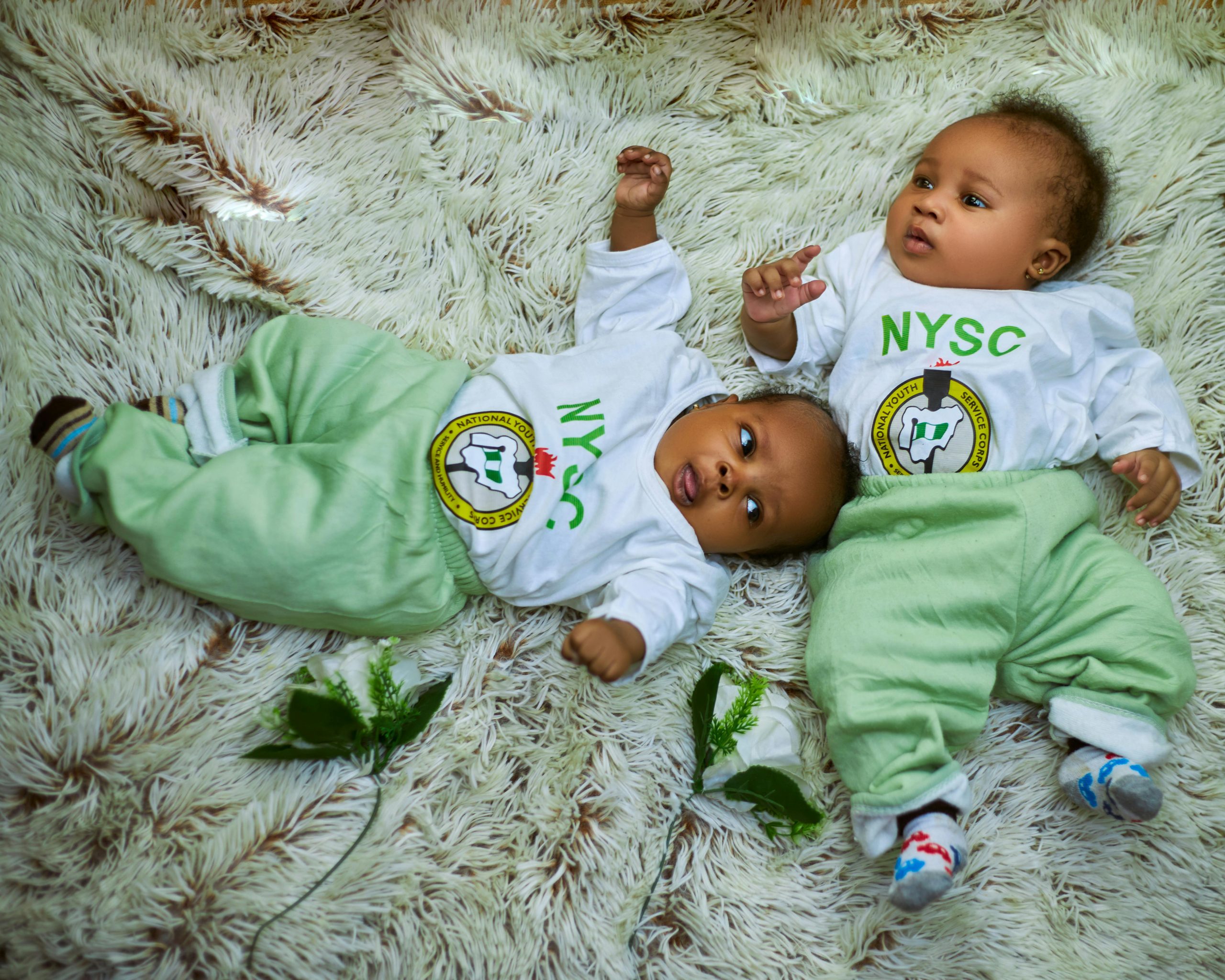 Adorable babies in NYSC attire lying on a cozy rug, representing innocence and national pride.